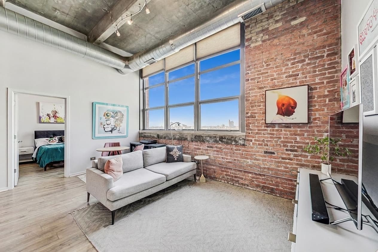 Living area with exposed brick, tall windows, and a peek into bedroom two